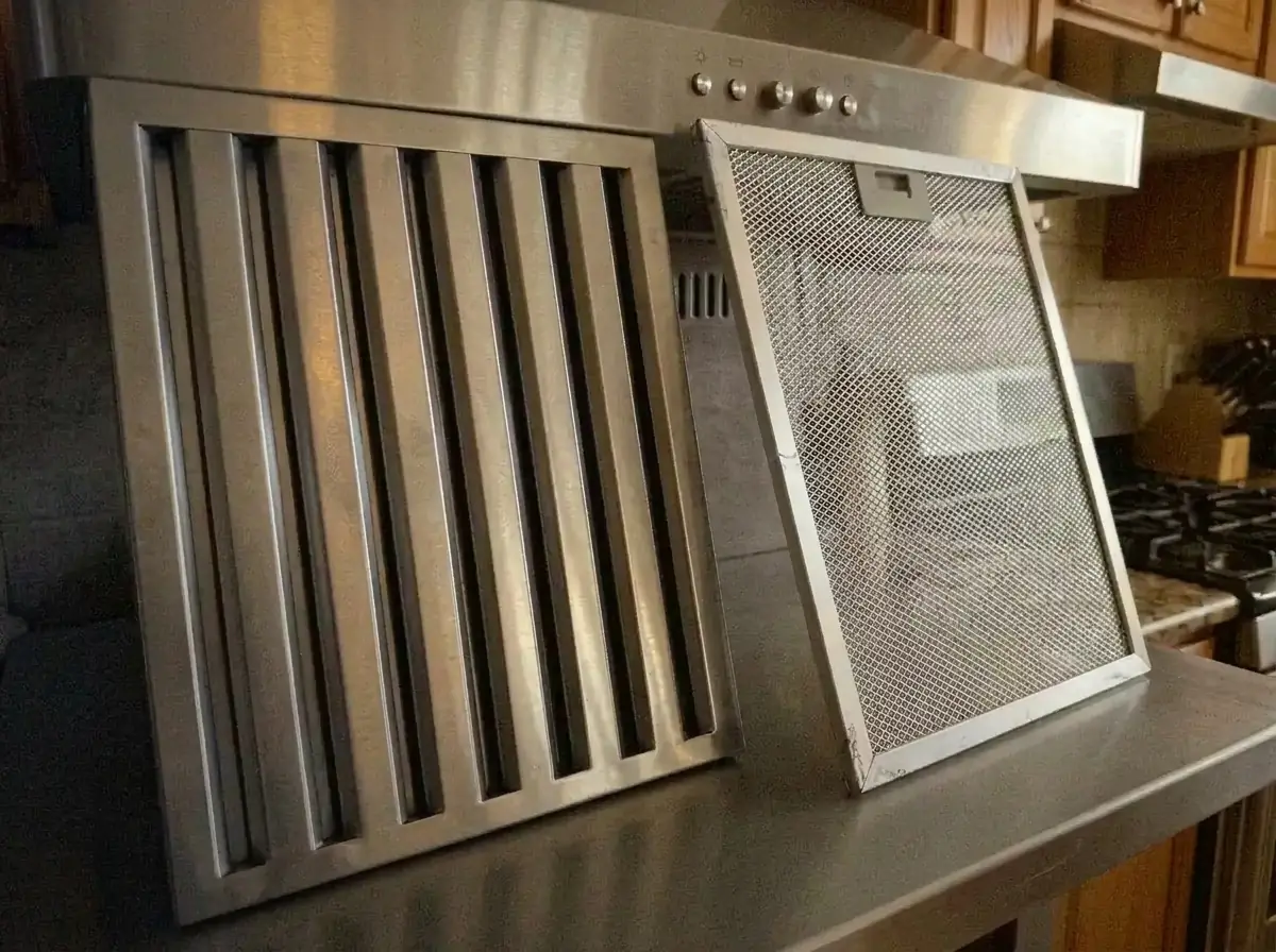 Stainless steel baffle grease filter and aluminum mesh grease filter side by side against a range hood, showing the two most common range hood filter types